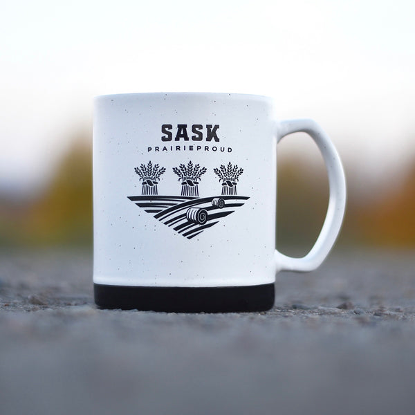 A ceramic mug with a white body and black base, featuring the text 'SASK Prairie Proud' and a graphic of wheat.