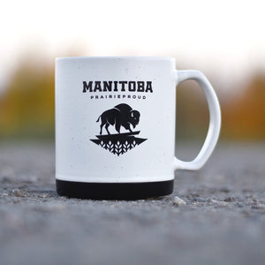 A ceramic mug with a white body and black base, featuring the text 'MANITOBA' and a buffalo silhouette, placed on a surface with a blurred natural background.
