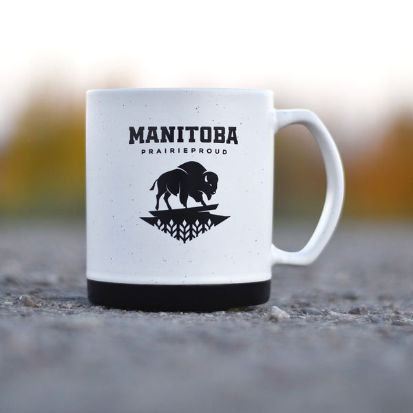 A ceramic mug with a white body and black base, featuring the text 'MANITOBA' and a buffalo silhouette, placed on a surface with a blurred natural background.