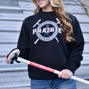 A person wearing a black crew neck sweater with the text 'Prairie Proud' and a curling inspired design.