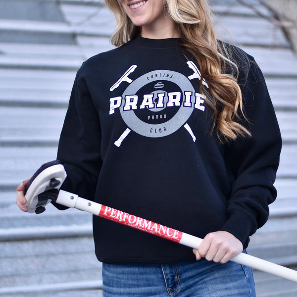 A person wearing a black crew neck sweater with the text 'Prairie Proud' and a curling inspired design.