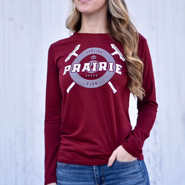 A female model wearing a long-sleeved red t-shirt with 'Prairie Proud' text and curling stone graphics on it, paired with jeans.