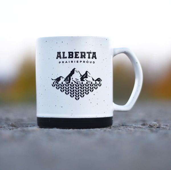 A ceramic mug with 'Alberta Prairie Proud' printed on it, predominantly white with a black base and black speckles, set against an outdoor backdrop.