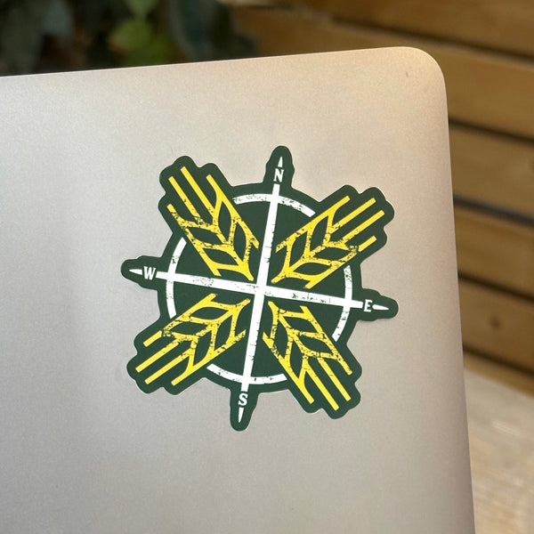 A die cut vinyl sticker with a compass design and wheat sheafs in forest green and gold colours, placed on a laptop.