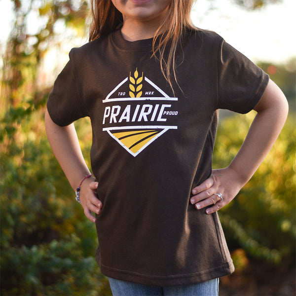 A child wearing a brown crew t-shirt with 'PRAIRIE PROUD' text and a graphic on it, standing outdoors.