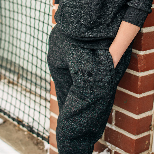 Youth wearing heather charcoal sweatpants with embroidered icons on the left hip, standing next to a brick wall.