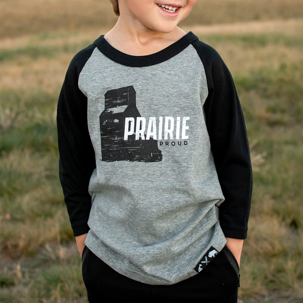 A child wearing a heather grey and black 3/4 raglan t-shirt with the text 'PRAIRIE PROUD' on the front.