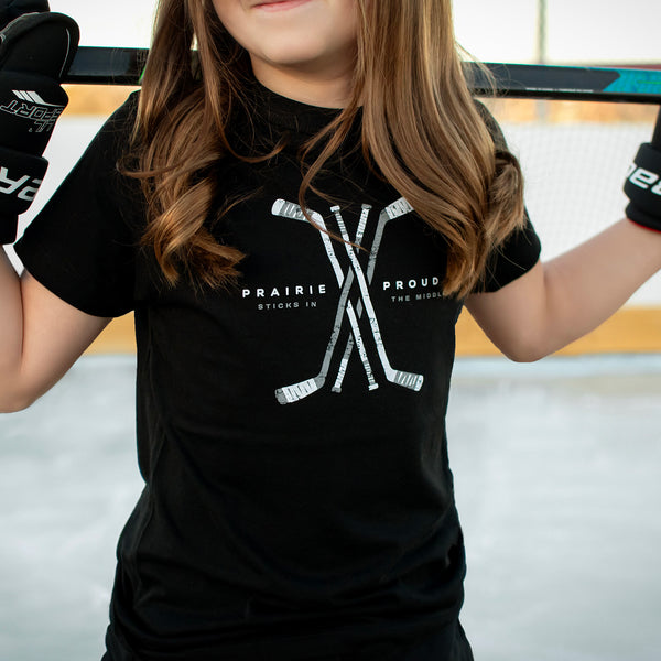 A child wearing a black crew neck t-shirt with 'PRAIRIE PROUD' and hockey stick graphics on it, standing on an ice rink.