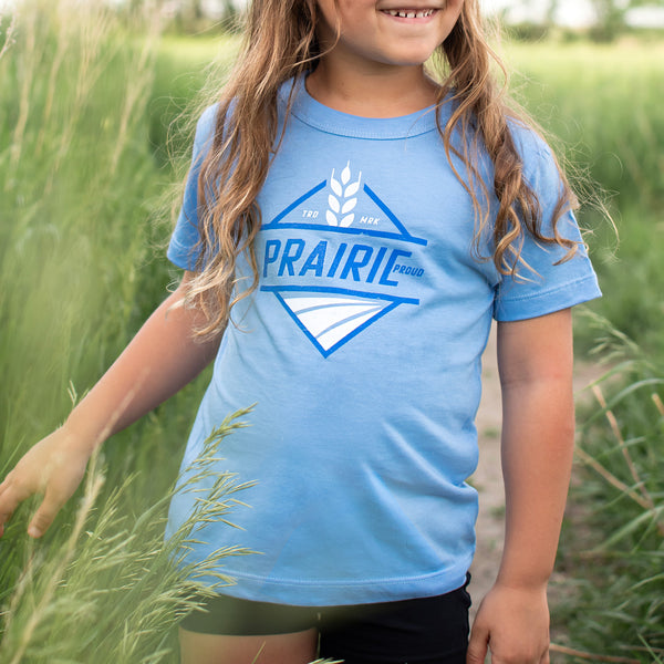 A child wearing a light blue crew t-shirt with the text 'PRAIRIE PROUD' printed on the front, standing in a field.