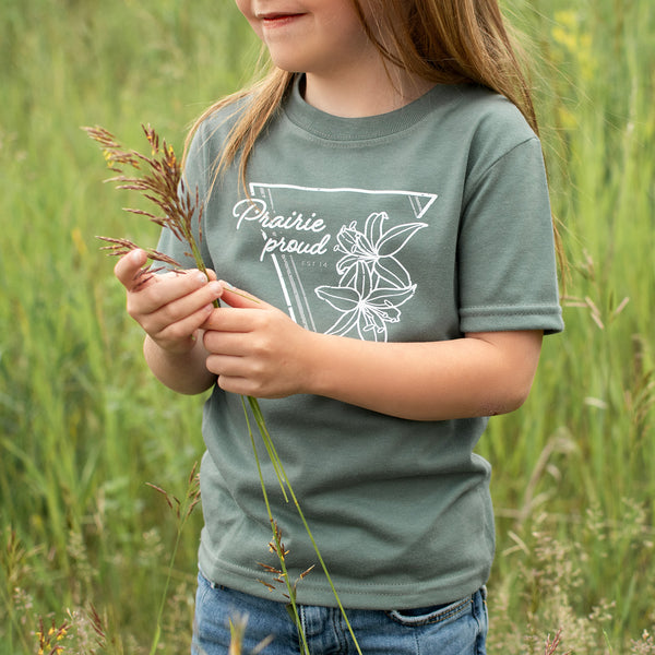 A child standing in a field holding grass, wearing a dusty green crew t-shirt with text and a flower design.