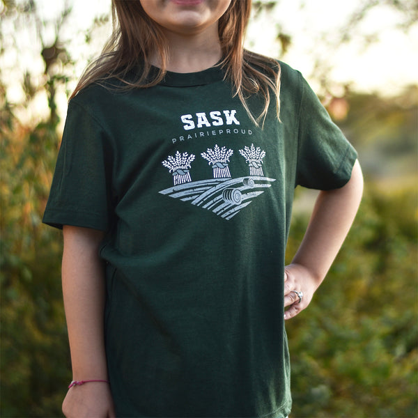 A child wearing a green crew t-shirt with 'SASK' text and a graphic on it, standing outdoors.