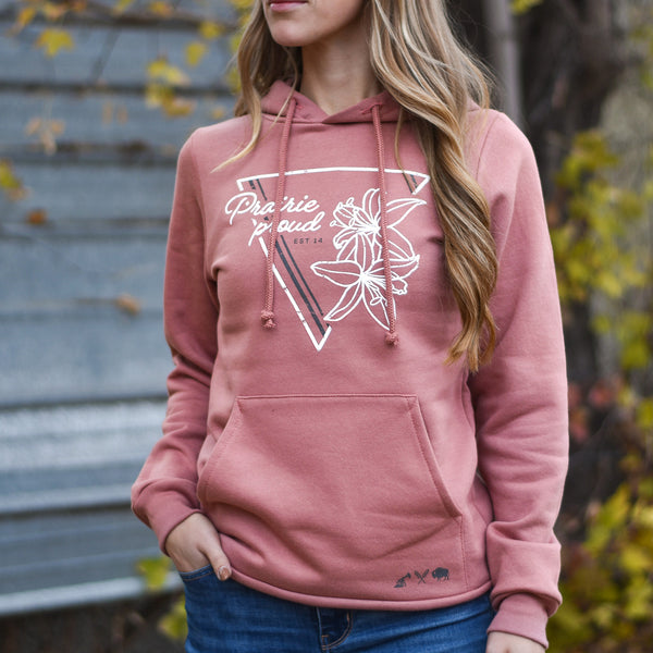 A woman wearing a dusty rose coloured hoodie with a triangular lily flower design and text 'prairie proud' on the front.