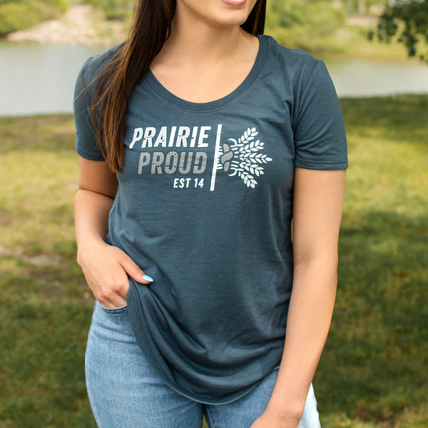 A woman wearing a blue scoop neck t-shirt with the text 'PRAIRIE PROUD' printed on the front next to a bundle of wheat.