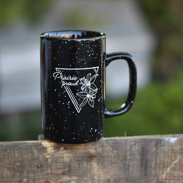 A black ceramic mug with white speckles and a floral design, placed on a wooden surface with a blurred background.