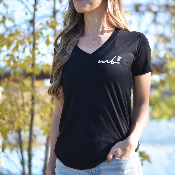 A woman standing outdoors wearing a black V-neck T-shirt with the text 'mb' (Manitoba) on the top left side.