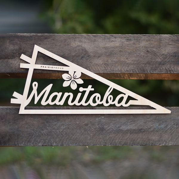 A wooden pennant with the text 'Manitoba' on it, placed on a wooden surface with foliage in the background.