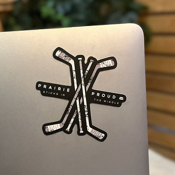 A black and grey die cut vinyl sticker with the phrases 'Sticks in the middle' and 'Prairie Proud' written on it with crossed hockey sticks in the centre.