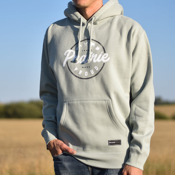 A person wearing a dusty mint green hooded sweatshirt with a circle design and the text 'prairie proud'.