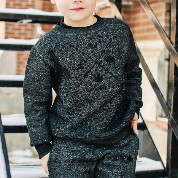 A child wearing a heather charcoal crew sweater with front embroidery, standing in front of a staircase.