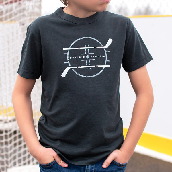 A child wearing a charcoal-colored T-shirt with a hockey-inspired design, standing near a hockey rink.