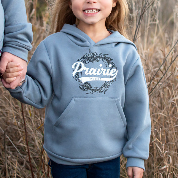 A child wearing a dusty blue hooded sweatshirt with the text 'Prairie Proud' on it, standing in a field of dry grasses.