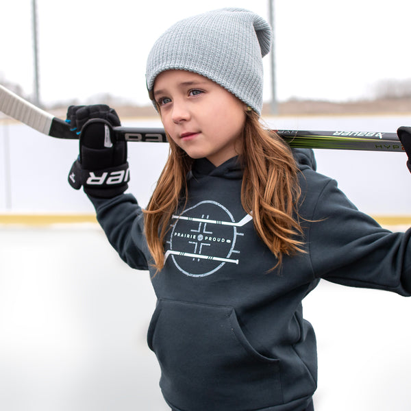 A child on an ice hockey rink wearing a gray hoodie with a hockey-themed graphic, a gray beanie, and holding a hockey stick.