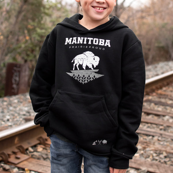 A child wearing a black hoodie with 'Manitoba' text and a bison graphic on it, standing by railway tracks.