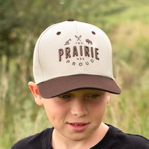 Child wearing a brown & cream coloured cap with 'Prairie Proud' text in an outdoor setting