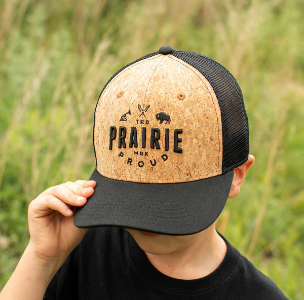Child holding a cork and black trucker hat on their head with the text 'Prairie Proud' against a grassy background