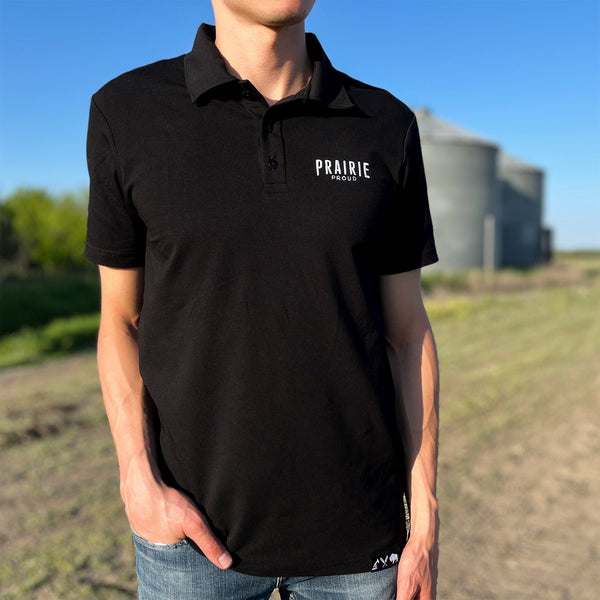 A male model wearing a black athletic polo shirt with the text 'Prairie Proud' embroidered on the left chest area, standing outdoors.