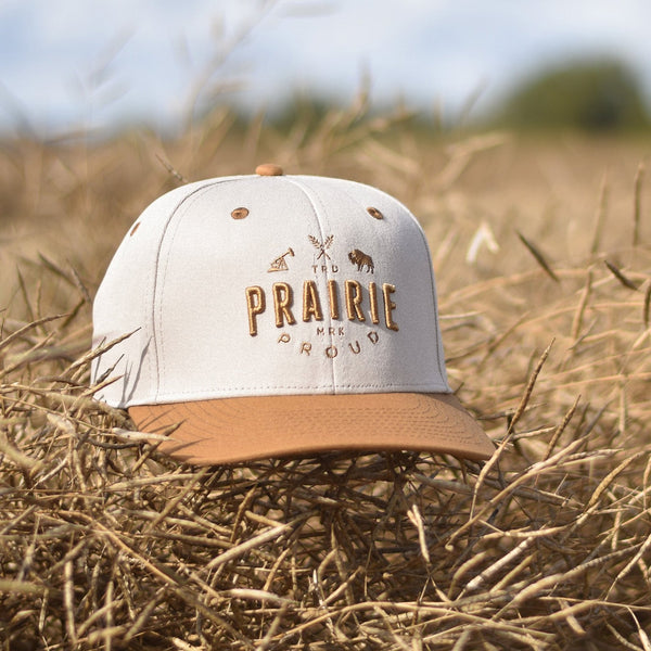 A snapback hat with a caramel coloured brim and a white crown, with caramel coloured text that says 'Prairie Proud'.