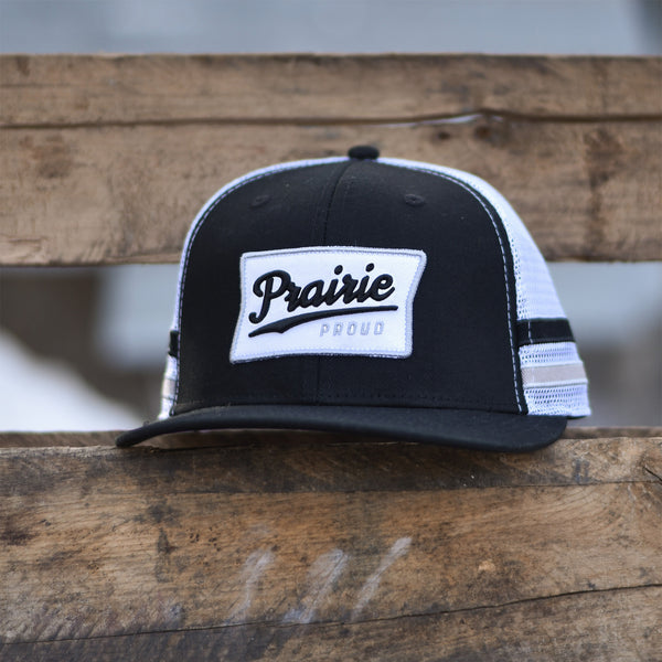 A black and white trucker hat with 'Prairie Proud' written on the front.