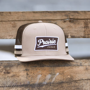 A brown, white, and tan trucker hat with 'Prairie Proud' written on the front.