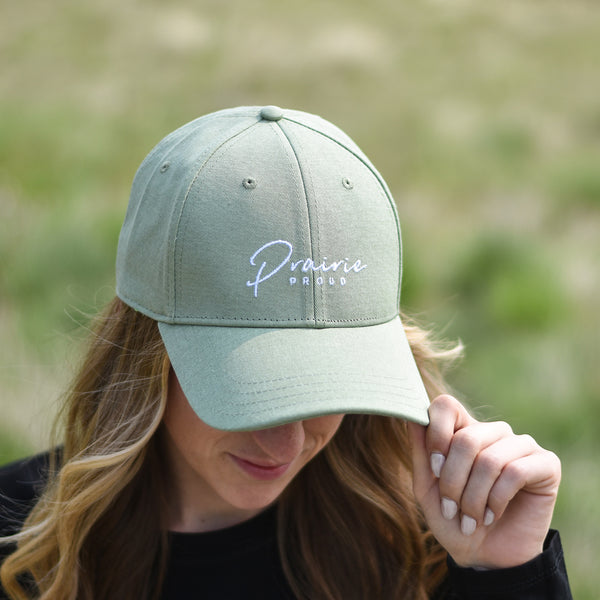 A person wearing a light green baseball cap with an adjustable slider, featuring a subtle logo with text that says 'prairie proud'.