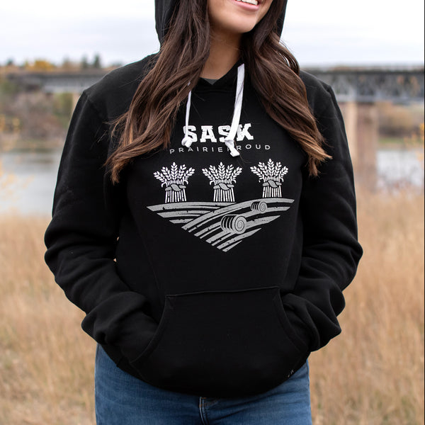 A person wearing a black hoodie with 'SASK' written on it with wheat in a field.