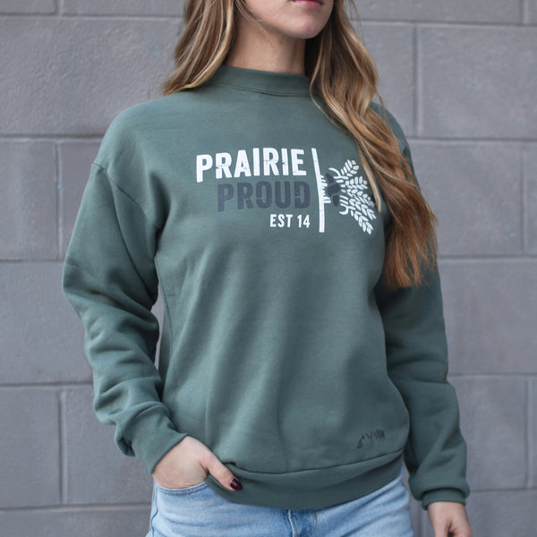 A person wearing a dusty green drop shoulder sweater with the text 'PRAIRIE PROUD EST 14' on the front.