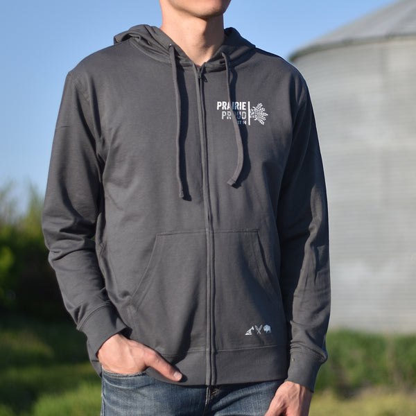 A person wearing a gray zip hoodie with a logo on the chest, standing outdoors.