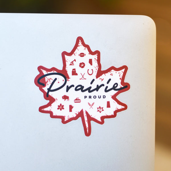 A red and white sticker in the shape of a maple leaf with various icons inside and 'Prairie Proud' written on it, placed on a laptop.