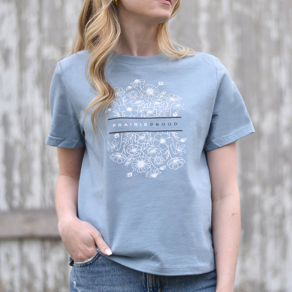 A woman wearing a dusty blue crew neck t-shirt with a floral design and the text 'PRAIRIE PROUD' on the front.