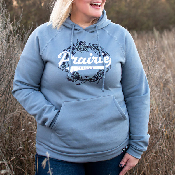 A woman wearing a dusty blue hooded sweatshirt with the text 'Prairie proud' on it, standing in a field.