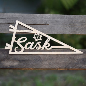 A wooden pennant with the text 'Sask' on it, placed on a wooden surface.
