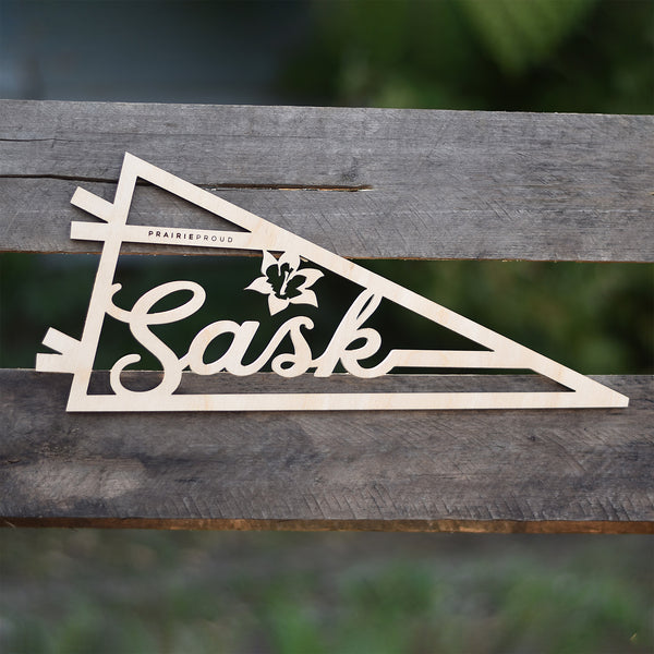 A wooden pennant with the text 'Sask' on it, placed on a wooden surface.