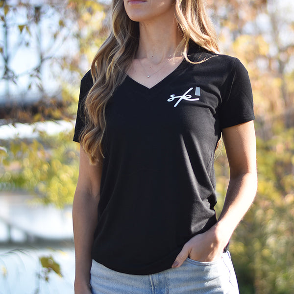 A woman standing outdoors wearing a black V-neck t-shirt with the text 'sk' (Saskatchewan) on the top left side.
