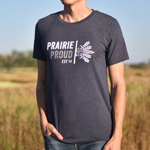 A person wearing a heather midnight blue crew t-shirt with the text 'PRAIRIE PROUD' and a graphic on it standing in a field.