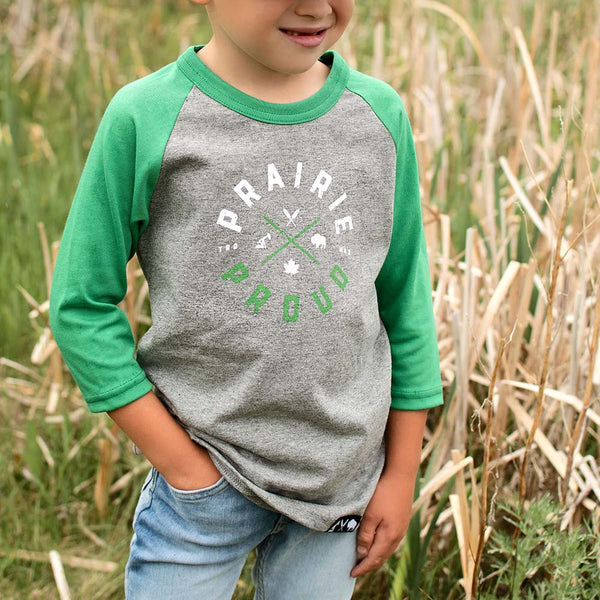 A child wearing a heather grey and green raglan t-shirt with the text 'PRAIRIE PROUD' on it, standing in a field.