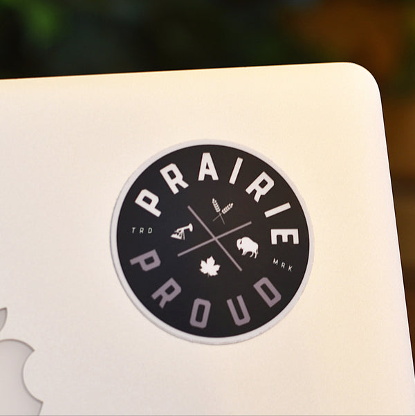 A die cut vinyl sticker with 'Prairie Proud' text and various symbols inside the circle, placed on a laptop.