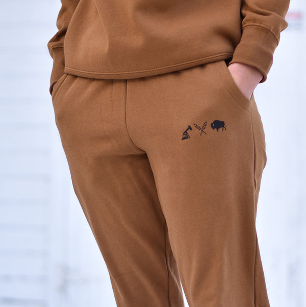 A person wearing caramel-coloured unisex joggers with a small black logo on the left leg.