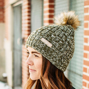 A person wearing a heather green and white cable knit beanie with a brown pom-pom on top.