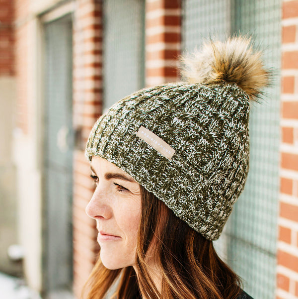 A person wearing a heather green and white cable knit beanie with a brown pom-pom on top.
