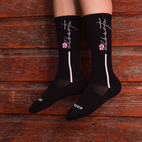 A pair of black crew socks with white text 'alberta' and pink flower on the ankle area, displayed against a wooden background.
